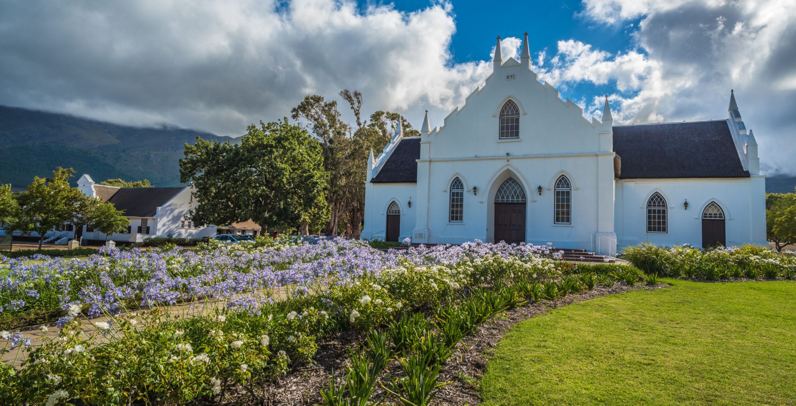 En smuk, hvid bygning med blomsterbede foran og en dramatisk himmel i Franschhoek