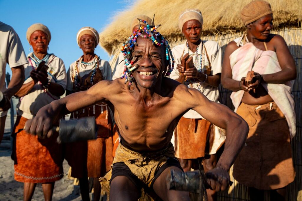 Khwai-landsbyboere står i en halvcirkel med en mand i forgrunden, der danser og smiler.