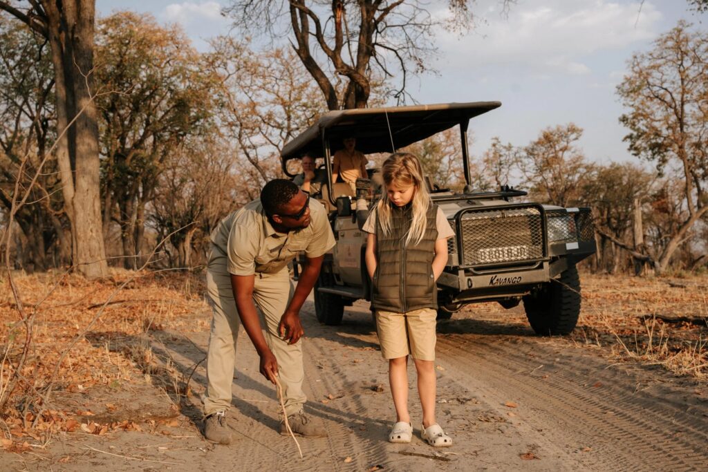 En mand viser dyrespor på den støvede vej til en ung pige med en safarijeep i baggrunden.